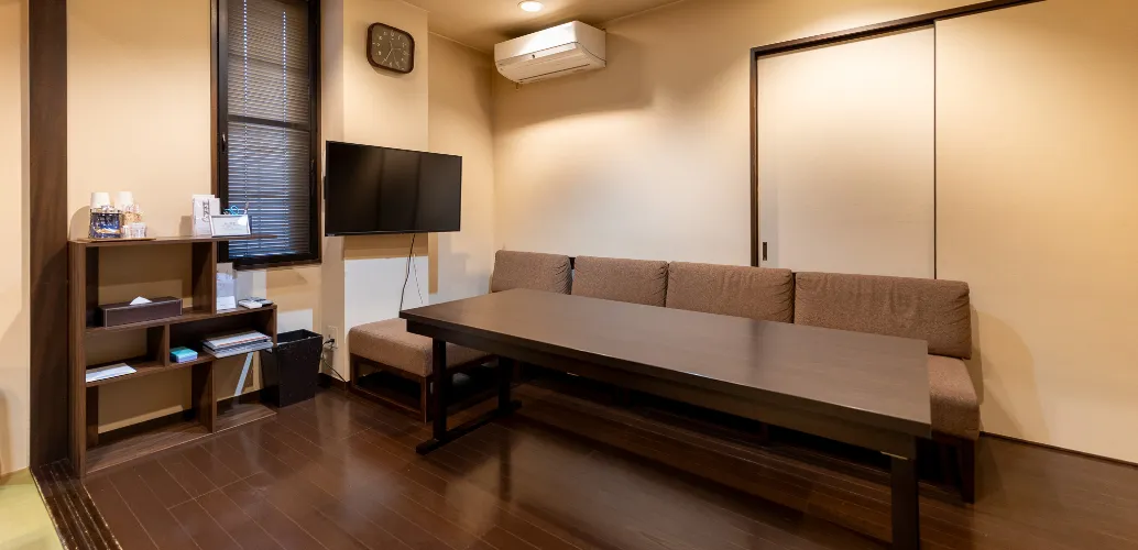 写真：エアコン、テレビ、ソファ、長テーブル、壁沿いに物がおける棚があるリビング